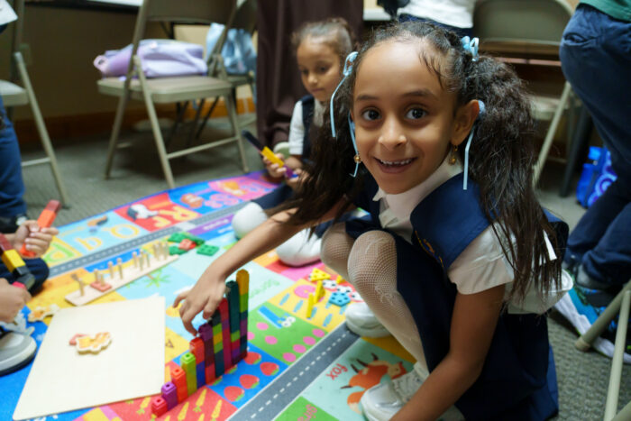 A smiling student stacks colorful blocks on a play mat as other children play in the background.