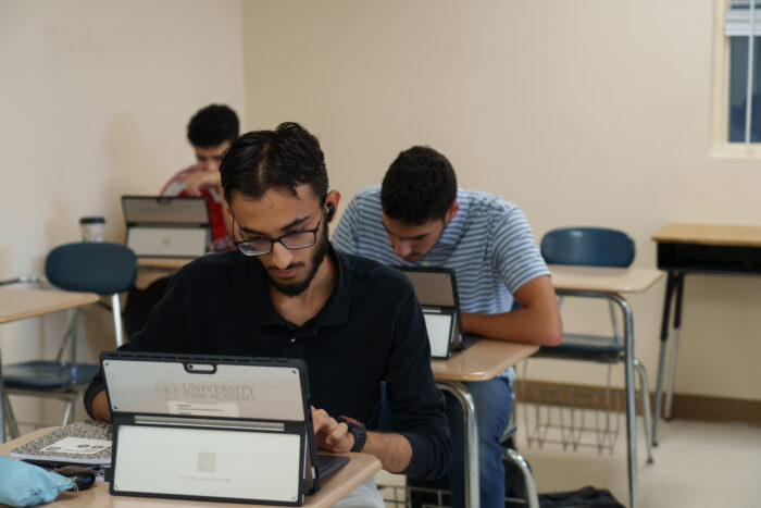 Three students in a classroom sit at desks using laptops.