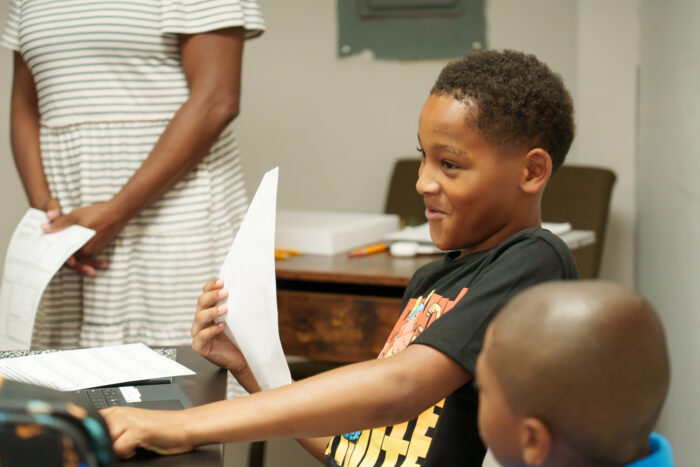 A smiling student holds paper at a table with another child.