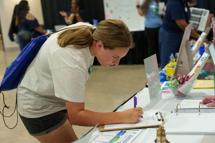 A student signs a paper while leaning over a table at open house.