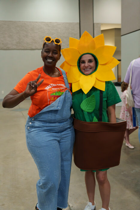 Two staff members pose indoors: one in overalls and an orange shirt, the other in a sunflower costume with a brown pot.