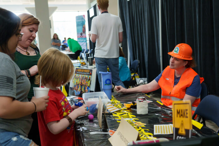 A student talks to a teacher who is seated at a table and dressed in a construction worker costume.