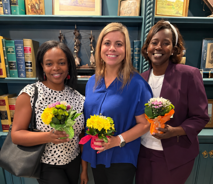 Three principals stand in a library, each holding a small bouquet of yellow and white flowers.
