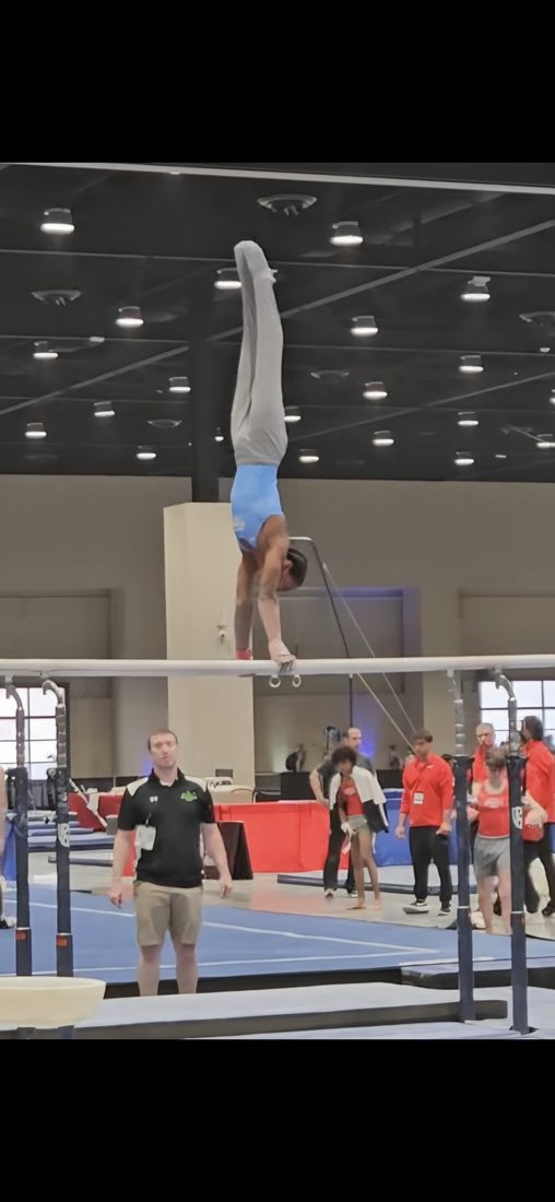 Josiah J does a handstand on parallel bars as a coach watches from below.