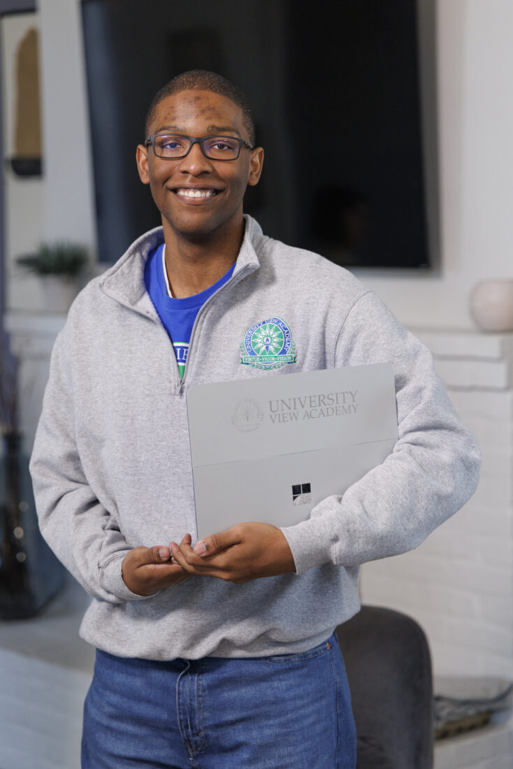 A smiling student in a University View Academy sweatshirt holds a branded laptop.