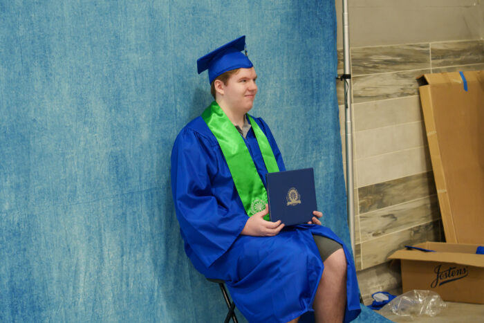 A student in a blue cap and graduation gown with a green stole holds a diploma and poses for a picture.