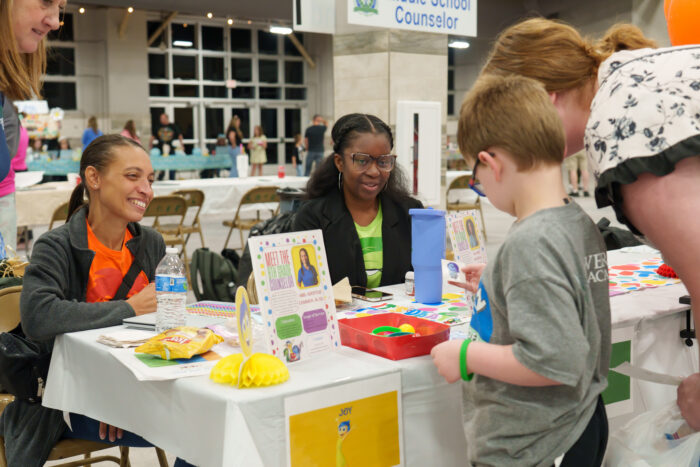 Two seated staff members smile at a student completing an activity at their table.