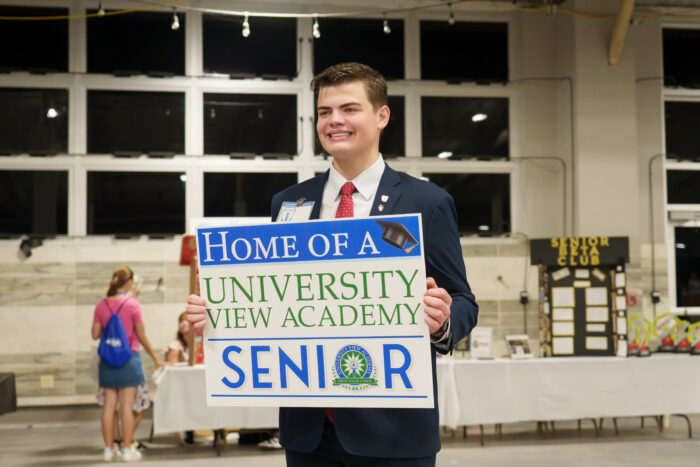 A student in a suit holds a sign reading, “Home of a University View Academy Senior.”.