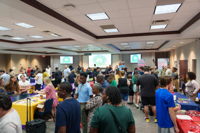 A wide photo of a large room filled with tables and several people walking around.