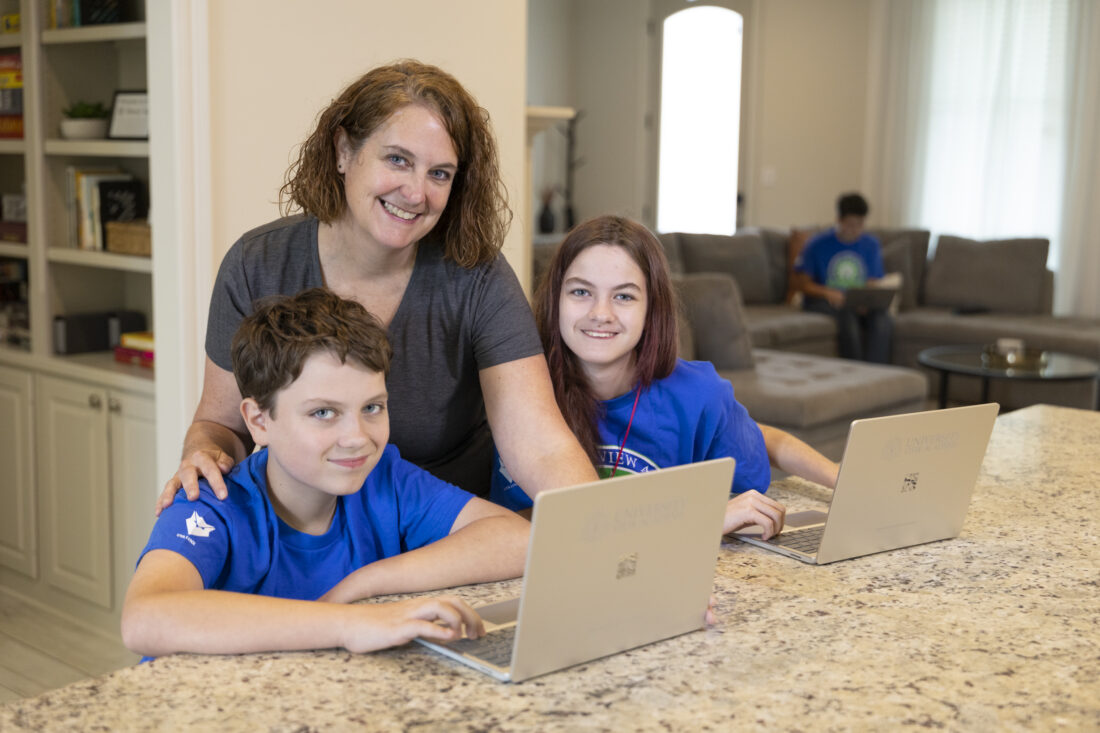 A woman and two children use laptops at a kitchen island, while another person sits on a sofa in the background.