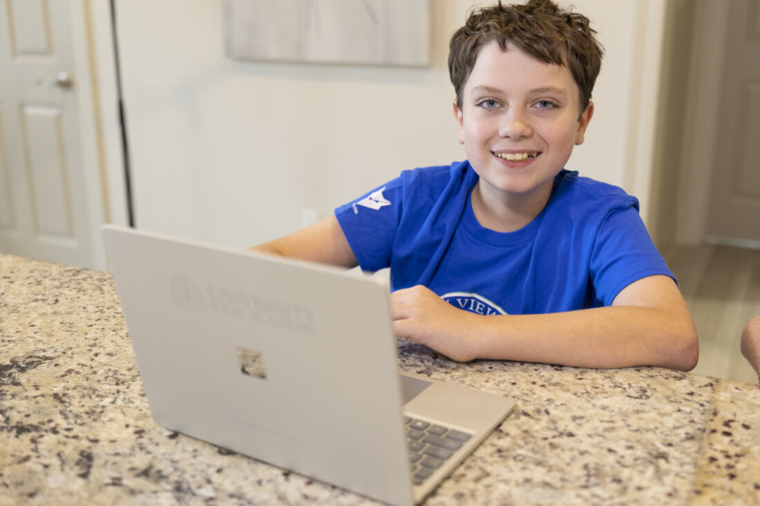 A smiling boy in a blue t-shirt sits at a kitchen counter with a laptop.