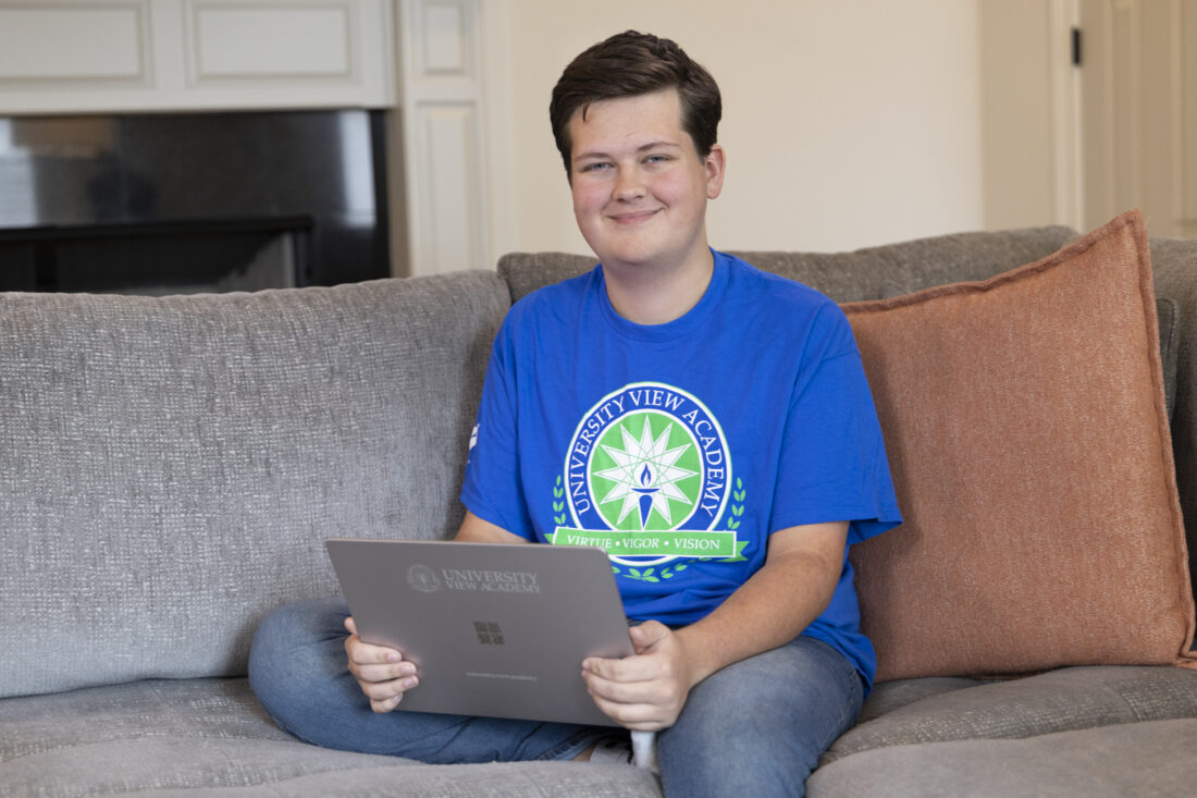 A young man in a University View Academy t-shirt sits on a sofa holding a matching laptop.