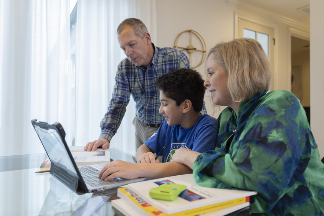 A boy sits at a table using a laptop, with two adults looking on and assisting with his schoolwork.